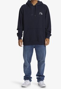 Sudadera con capucha azul marino con bolsillo canguro y logo blanco en el pecho, combinada con vaqueros azules y zapatillas blancas. Tela suave, ajuste relajado.