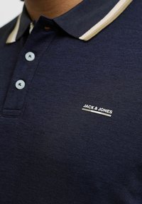 Tums zils polo krekls ar bēšu un baltu svītru apkakli, gaiši ziliem pogām, ar baltu "JACK & JONES" logotipu uz krūtīm.