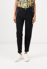 Žena v černých skinny džínách s vysokým pasem, doplněná bílými teniskami a květinovou blůzkou s krátkým rukávem.