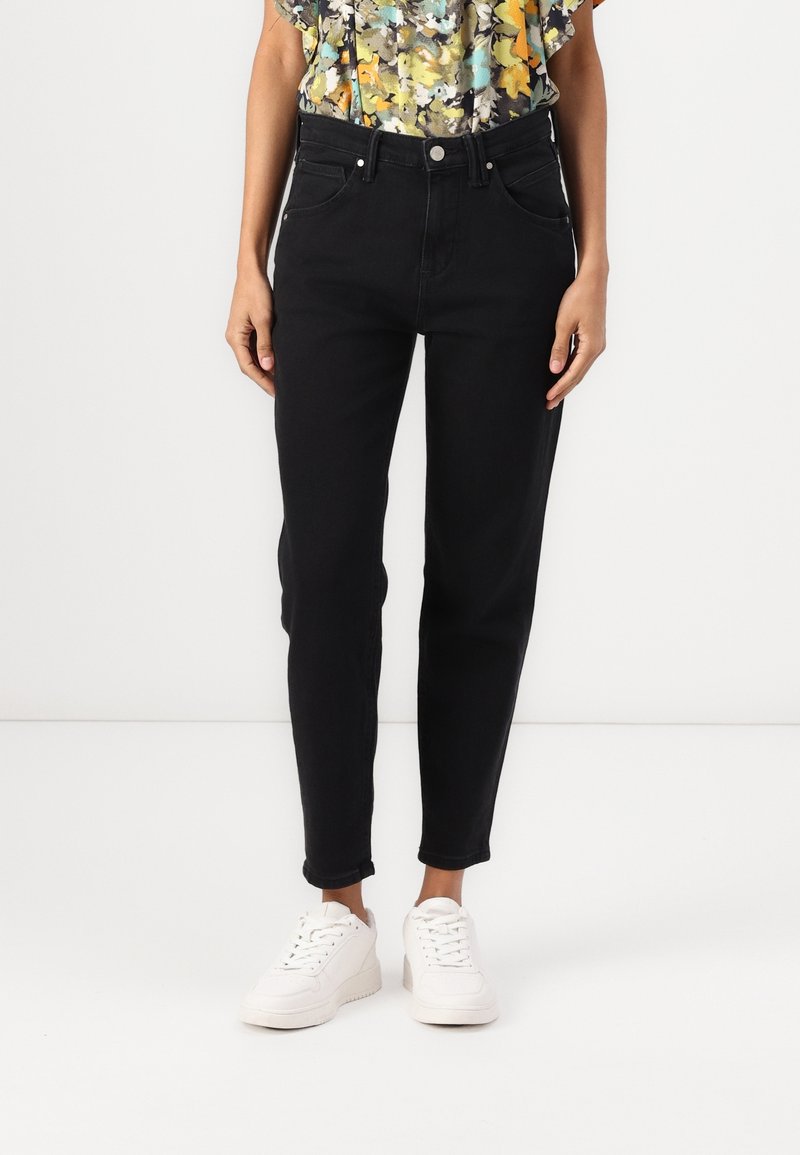 Žena v černých skinny džínách s vysokým pasem, doplněná bílými teniskami a květinovou blůzkou s krátkým rukávem.