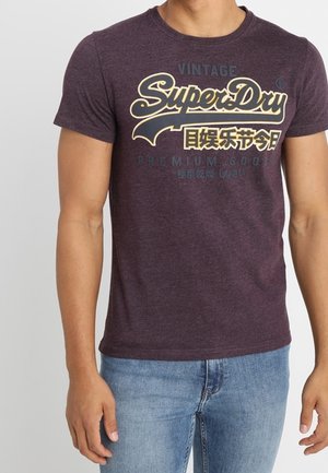 Mężczyzna w ciemnofioletowej koszulce SuperDry z żółtym logo i spranych dżinsach, stojący na tle jasnego, jednolitego tła.