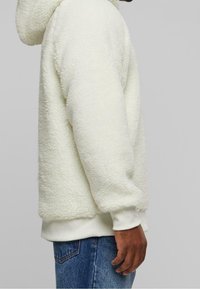 Felpa con cappuccio color crema in pile con superficie morbida e testurizzata, polsini e fascia in vita a coste. Il design comprende un cappuccio e dettagli sulle cuciture laterali.