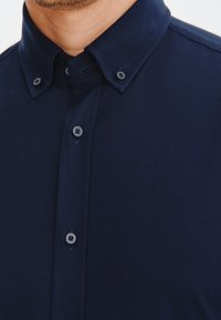 Koszula w kolorze granatowym, zapinana na guziki, wykonana z teksturowanej tkaniny, z ostrym kołnierzykiem i kontrastującymi guzikami. Posiada czysty, dopasowany krój.