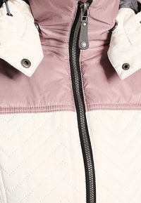 Veste matelassée à motifs en losanges avec une section supérieure rose clair, une section inférieure crème, une couture diagonale et une fermeture éclair centrale avec boutons-pression.