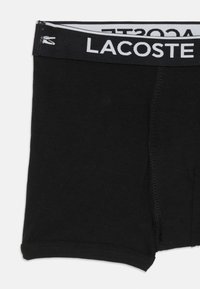 Juodos medvilninės kelnaitės su vientisu dizainu, turinčios juodai baltą prekės ženklo juostą, pažymėtą "LACOSTE." Minkšta tekstūra, vidutinio ilgio kojos.