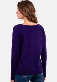 Lila Pullover mit langen Ärmeln, Rundhalsausschnitt und weicher Textur. Formgeschnittener Saum auf der Rückseite, kombiniert mit blauen Jeans.