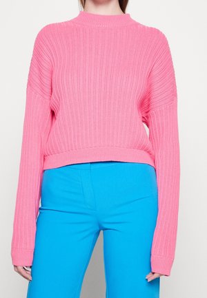 Personne portant un pull rose côtelé, ample, à manches longues, et un pantalon bleu vif taille haute, se tenant devant un fond uni.