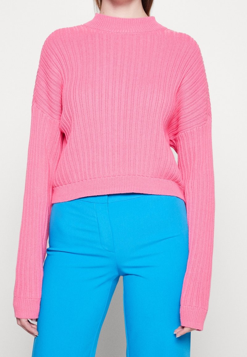 Personne portant un pull rose côtelé, ample, à manches longues, et un pantalon bleu vif taille haute, se tenant devant un fond uni.