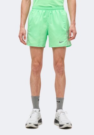 Uomo che indossa pantaloncini da corsa Nike verde brillante, calzini Nike grigi e scarpe da ginnastica Nike bianche, in piedi davanti a uno sfondo semplice.