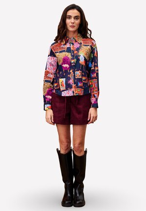 Femme portant une chemise à manches longues à motifs colorés, un short bordeaux et des bottes noirs jusqu'au genou, se tenant avec les bras détendus le long du corps.