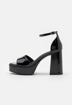 Sandales à plateforme en cuir verni noir avec un bout ouvert à l'avant et une bride autour de la cheville, présentant un épais talon carré et une texture lisse et brillante.