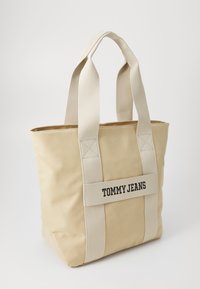 Bež kanvas torba s dva debela remena. Na prednjoj strani ima crni logo "TOMMY JEANS" na tkanom remenu. Jednostavan pravokutni dizajn.