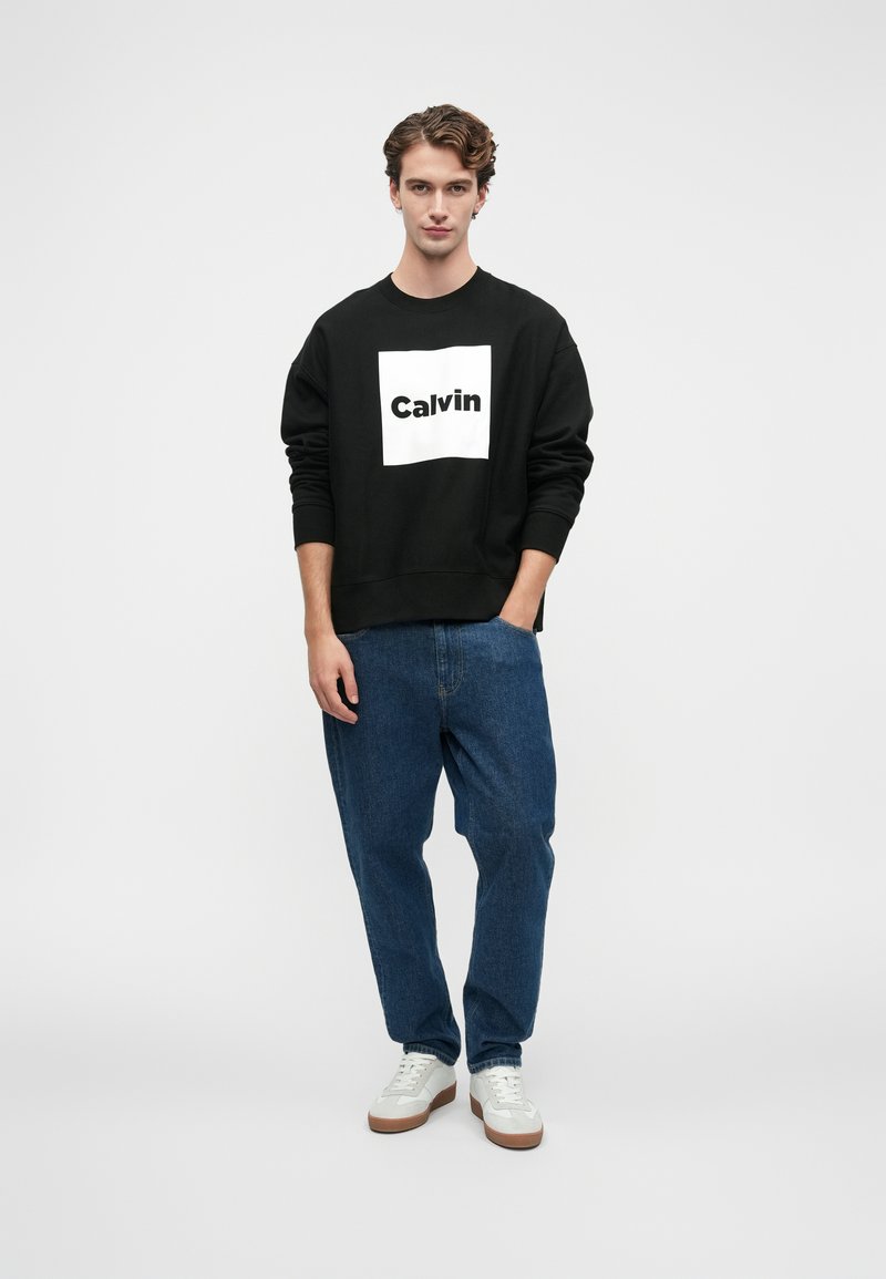 Mladý muž stojí s jednou rukou v vrecku, oblečený v čiernej mikine značky Calvin, modrých džínsoch a bielych teniskách na čistom bielom pozadí.