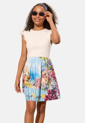 Meitene ar cirtainiem matiem, nēsā baltu bezroku kleitu ar krāsainu piejūras ciemata un ziedu rakstu svārkiem, regulē tumšās saulesbrilles.