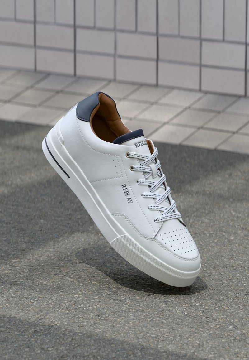 Weißer Sneaker mit niedrigem Schnitt und marineblauen Akzenten, perforierter Spitze, Schnürdesign und "REPLAY"-Markierung an der Seite und auf der Zunge, auf Asphalt platziert.
