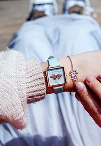 Montre à cadran carré avec un motif de papillon, cadre doré et bracelet bleu clair. Bracelet en argent avec accent floral porté par une main dans un pull en tricot.