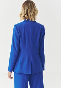 Modrý dámský blejzr na míru z hladké tkaniny. Má strukturovaný střih, výstřižky s klopami a výrazné zadní švy, které dodávají přiléhavý tvar.