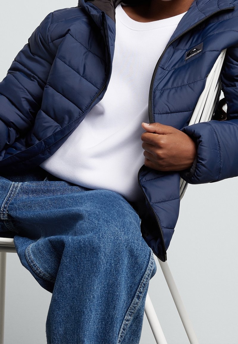Veste matelassée marine à capuche avec doublure intérieure noire et petit écusson logo, portée avec une chemise blanche et un jean bleu.