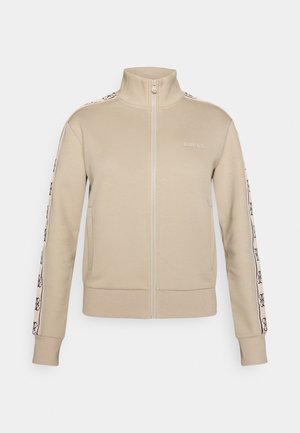 Beige jakke med glidelås laget av mykt materiale. Har høy krage, logo på brystet, og mønstrede sidelommer. Ribbestrikkede mansjetter og kanter.