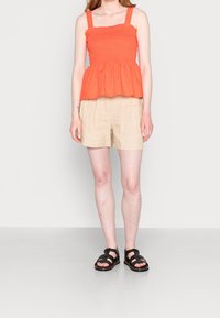 Top peplum di un brillante arancione con texture arricciata, abbinato a pantaloni corti di lino beige e sandali neri. Un outfit semplice e casual.