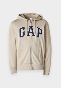 Sweat à capuche beige zippé avec lettrage "GAP" en bleu marine, capuche avec cordon, deux poches avant et poignets côtelés. Tissu doux avec texture lisse.