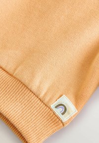 Angolo di maglione lavorato a maglia color pesca con orlo a costine e un'etichetta di tessuto con un contorno arcobaleno stilizzato in colori smorzati.