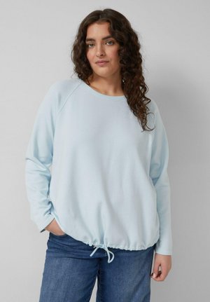 Kobieta z kręconymi ciemnymi włosami, ubrana w jasnoniebieską bluzę z kapturem i ściągaczem u dołu oraz niebieskie dżinsy, stojąca na tle jednolitego jasnoszarego tła.