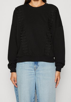 Schwarzer Pullover mit strukturierter Spitzenverzierung an den Schultern, gerippten Bündchen und Saum. Kombiniert mit hellblauen Jeans. Weicher Stoff und lockere Passform.