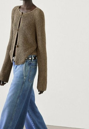 Person wearing a brown knit cardigan with large sleeves and loose blue jeans with a black belt, standing against a plain light wall.