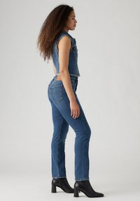 Chaleco corto de mezclilla azul con dobladillo deshilachado, combinado con jeans de pierna recta. La modelo lleva botas de tobillo con tacón bloque negro.