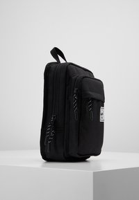 Czarny plecak o prostokątnym kształcie, wykonany z wytrzymałej tkaniny. Posiada wiele kieszeni na zamki oraz górny uchwyt do przenoszenia.