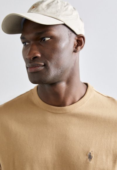 Baseball cap in light beige with a curved brim; tan crewneck shirt with subtle logo detail; smooth fabric texture.