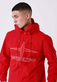 Jonge man met kort donker haar in een rode PXKD-jack met capuchon, kijkt naar links tegen een effen witte achtergrond.