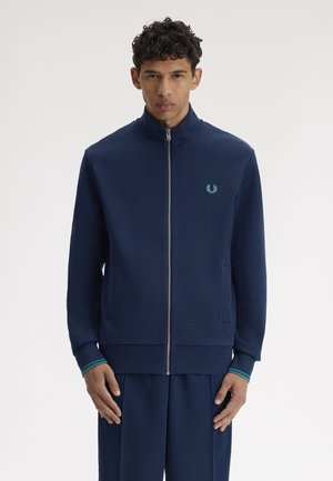 Mann trägt eine marineblaue Reißverschlussjacke mit hohem Kragen und Bündchen mit türkisfarbenen Streifen, steht vor einem weißen Hintergrund.