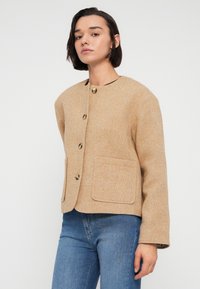 Μπεζ σακάκι από μάλλινο μείγμα με στρογγυλή λαιμόκοψη, τέσσερα μπροστινά κουμπιά, δύο εξωτερικές τσέπες και μακριά μανίκια. Λεία υφή, εφαρμοστή γραμμή.