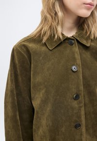 Ξανθός/ή φοράει ένα σουέτ παλτό σε ελιά πράσινο χρώμα με μαύρα κουμπιά, με γιακά ορατό, μπροστά σε μια απλή ανοιχτή φόντο.
