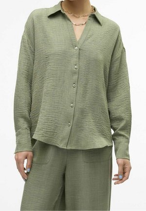 Femme portant une chemise boutonnée ample et texturée vert olive clair avec col et un pantalon large assorti, accessoirisée de colliers chaîne en or.