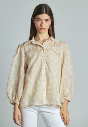 Blouse brodée beige clair avec un devant boutonné, un col et des manches bouffantes avec des poignets élastiques ; fabriquée à partir d'un tissu transparent et texturé.