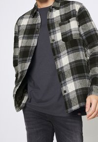 Rutigt skjorta i grå, svart och blå nyanser med knäppning och bröstficka. Lager på en mörk t-shirt och tillsammans med mörka jeans.