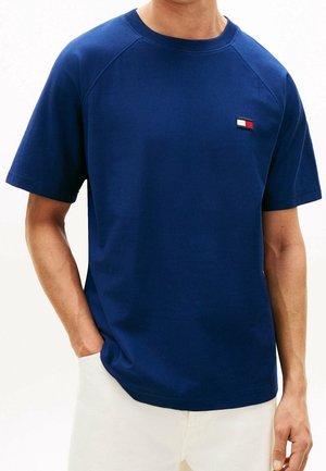 Homme portant un T-shirt bleu marine uni à manches courtes avec un petit logo rectangulaire rouge, blanc et noir sur la poitrine, associé à un pantalon de couleur claire.