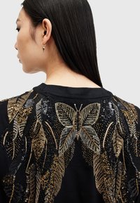 Svart hettegenser med intrikate detaljer i gull- og sølvpaljetter og broderi av blader og en sommerfugl på ryggen.