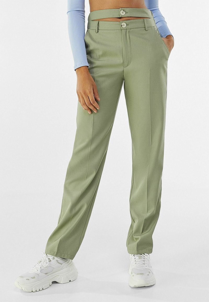 Pantalon vert clair ajusté avec une taille haute, deux poches avant, et une ceinture unique avec deux boutons. Porté avec des baskets blanches.