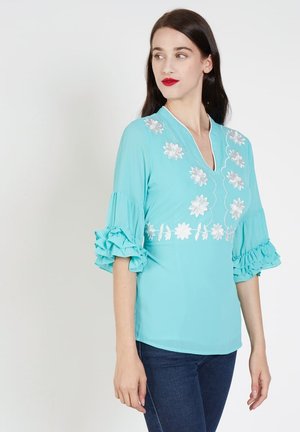 Blusa turquesa con mangas con volantes y bordado floral blanco en el escote y la cintura, hecha de un tejido ligero.