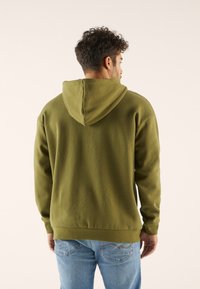 Olivgrüner Hoodie mit weicher Textur, Kängurutasche und angenattached Kapuze. Kombiniert mit hellblauen Jeans, die von hinten einen lässigen Schnitt zeigen.