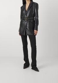 Zwarte leren blazer met een ceintuur in de taille, met afgeknipte revers en twee voorzakken, gestyled over aangesloten zwarte broek met uitlopende pijpen.
