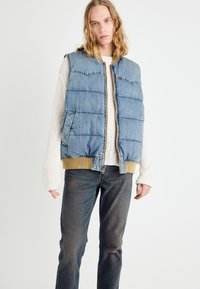 Lys blå denim puffer vest med glidelås foran, to sidelommer og ribbede mansjetter i sennepsgult. Båret over en kremfarget strikkegenser.