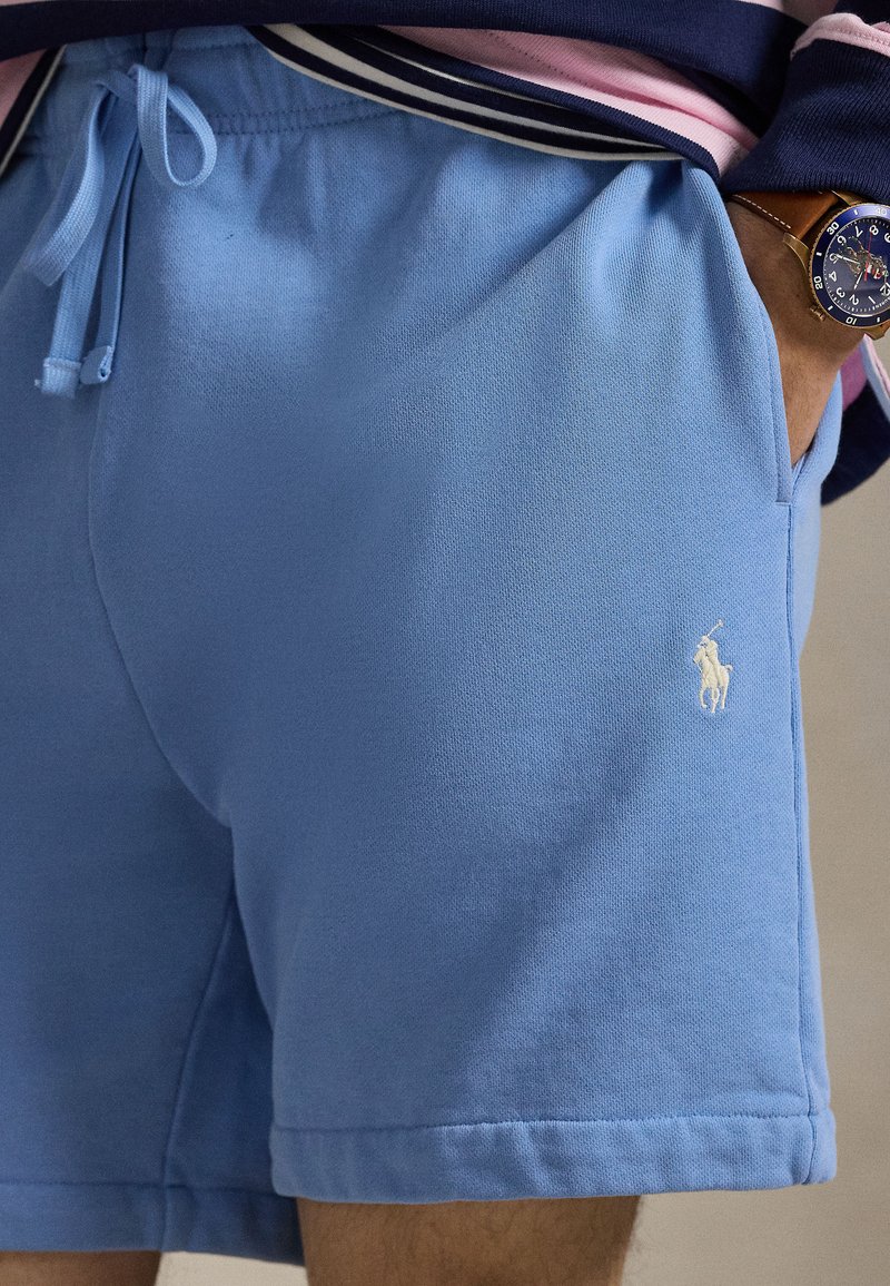 Man wearing light blue drawstring shorts with white embroidered horse logo and navy striped shirt, hand in pocket, watch on wrist.