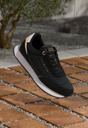 Black and gold Tommy Hilfiger sneaker with white sole on wooden block and gravel ground, with blurred greenery in the background.