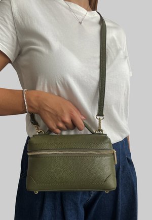 Borsa a tracolla in pelle verde oliva con superficie testurizzata, dotata di zip dorata e tracolla removibile, tenuta da una persona in una maglietta bianca.