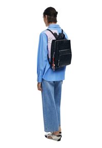 Person, die ein blau gestreiftes Hemd mit Cartoon-Grafiken, weite Jeans, weiße Sneaker und einen schwarzen, geprägten Rucksack mit Schlüsselanhänger-Zubehör trägt.